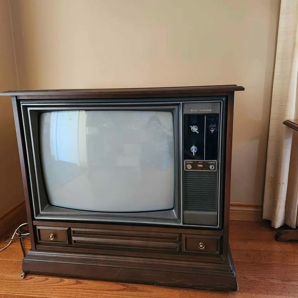 A vintage wooden-cased Electrohome television sits on a hardwood floor, its screen blank and reflective, showing a faint…