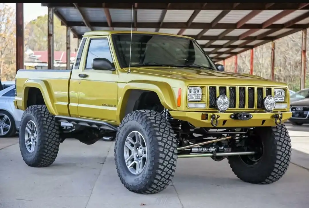 a yellow jeep comanche pickup truck is shown with a heavily modified lifted suspension, oversized knobby tires, and a custom…