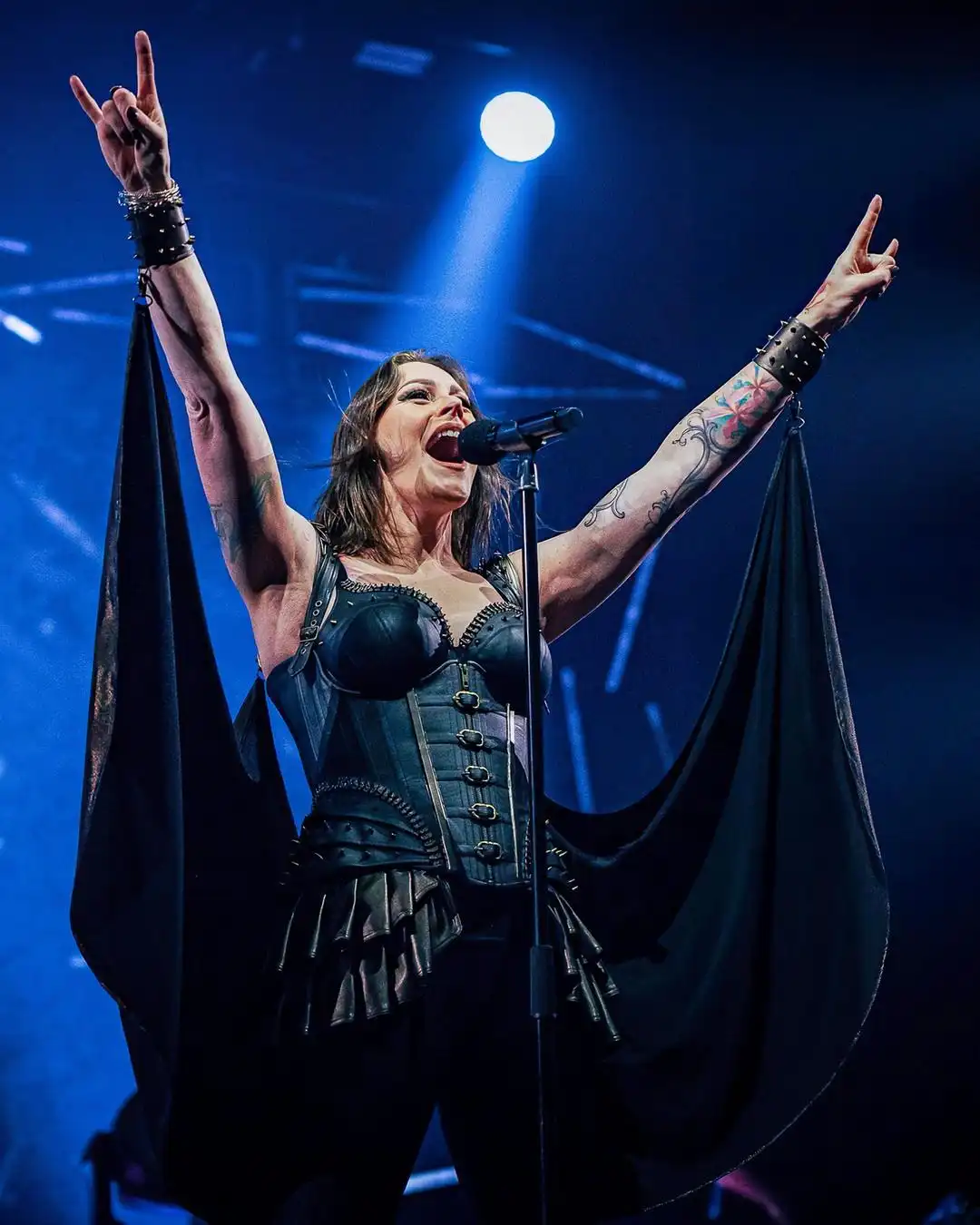 A female performer with tattoos and dark clothing, arms raised in a rock gesture, singing into a microphone under blue stage…