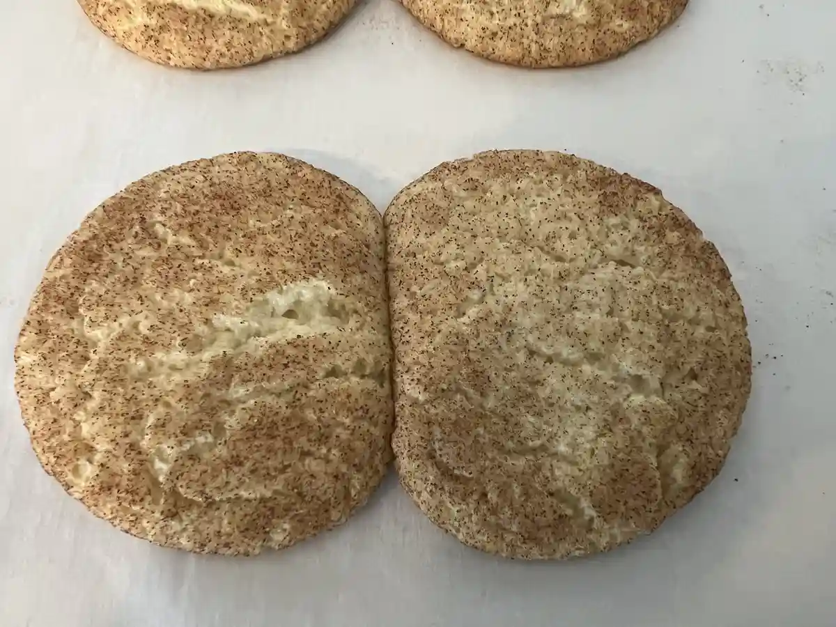 A close-up photo shows two halves of a round, golden-brown snickerdoodle cookie, split down the middle and placed…