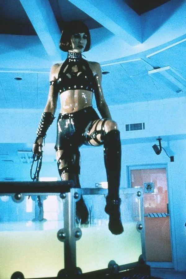 A woman in a shiny black latex outfit with a bob haircut stands on a transparent platform in a blue-lit room, holding a…