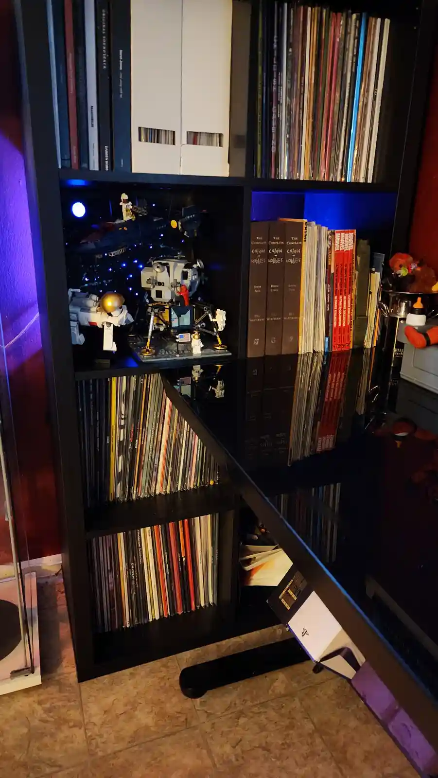 A black bookshelf filled with vinyl records, books, and a LEGO moon landing diorama with astronauts and a lunar module…