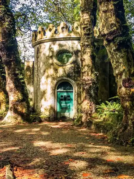 a small, round stone building with a teal door and crenellated roof sits nestled among large, mossy trees in a forest…