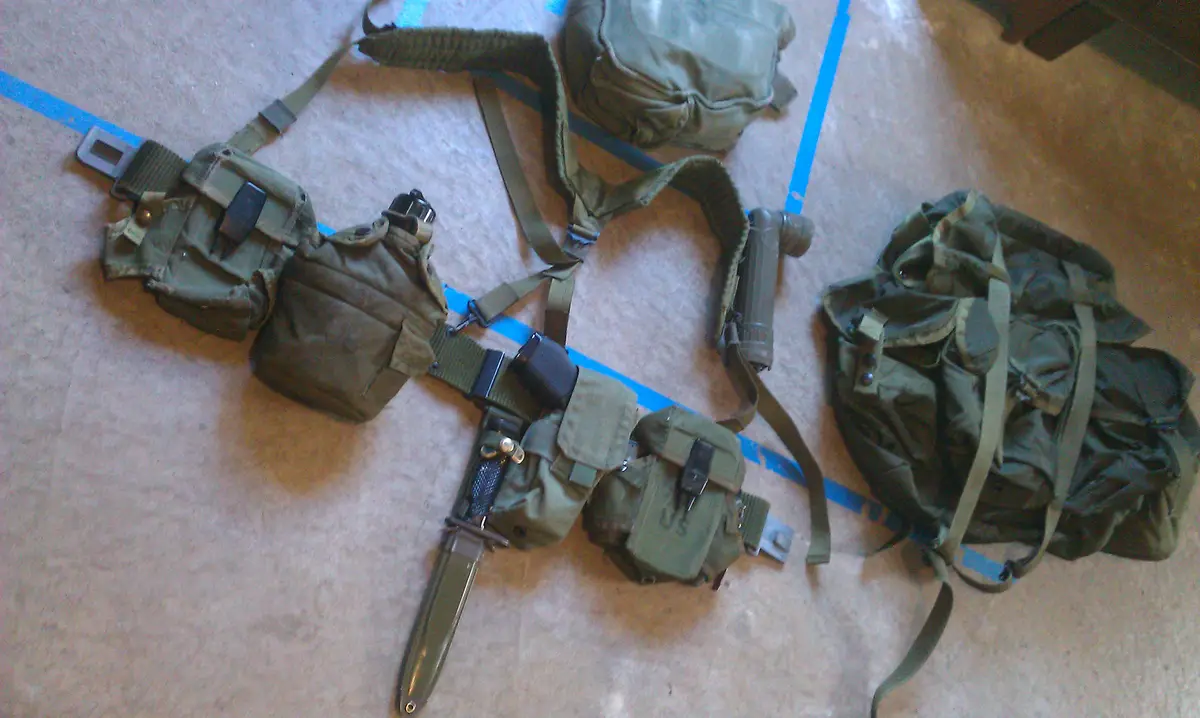A military-style gear setup, including a belt with pouches, a bayonet, and a backpack, is laid out on a carpeted floor…