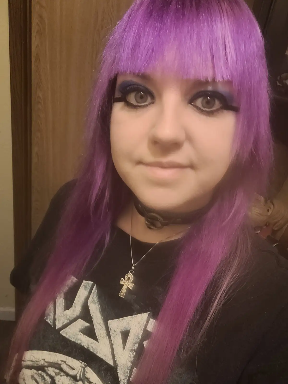 A person with vibrant purple hair and bangs, wearing heavy black eyeliner and a black t-shirt with a graphic, looks directly…