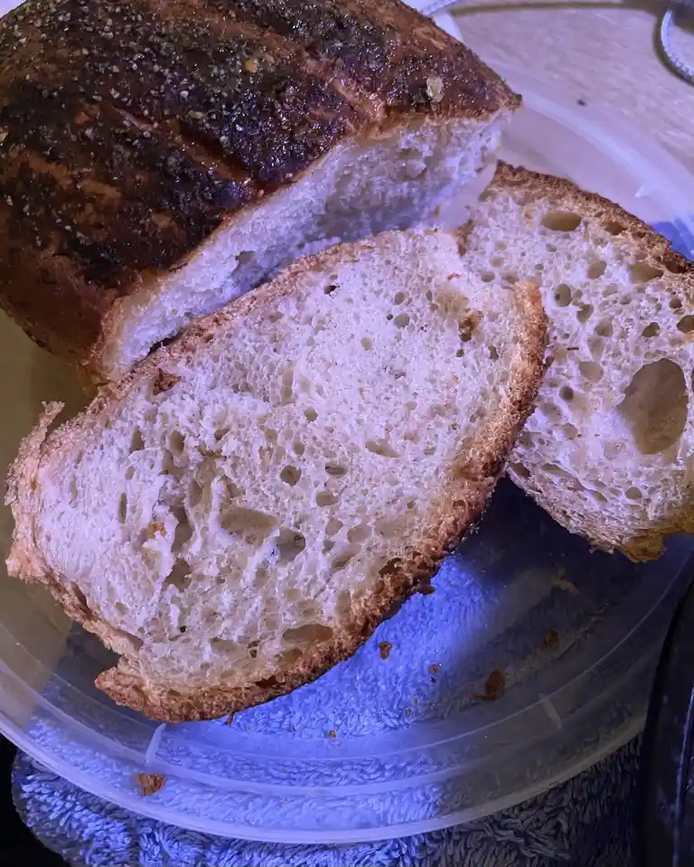 A close-up photo shows a rustic loaf of artisan bread with a dark, crackled crust and a soft, airy crumb, sliced open to…