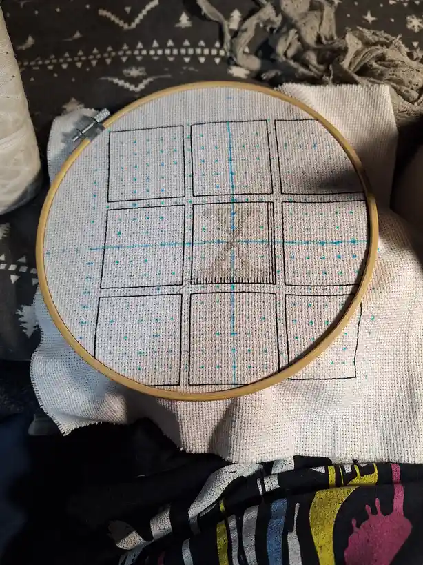 A close-up of a wooden embroidery hoop holding white fabric with a tic-tac-toe grid stitched in black thread. The center…