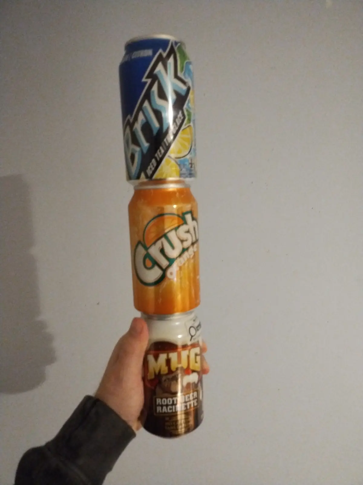 A hand holds a precarious tower of three soda cans against a plain wall: a blue 'Brisk Iced Tea Lemon' can on top, an orange…