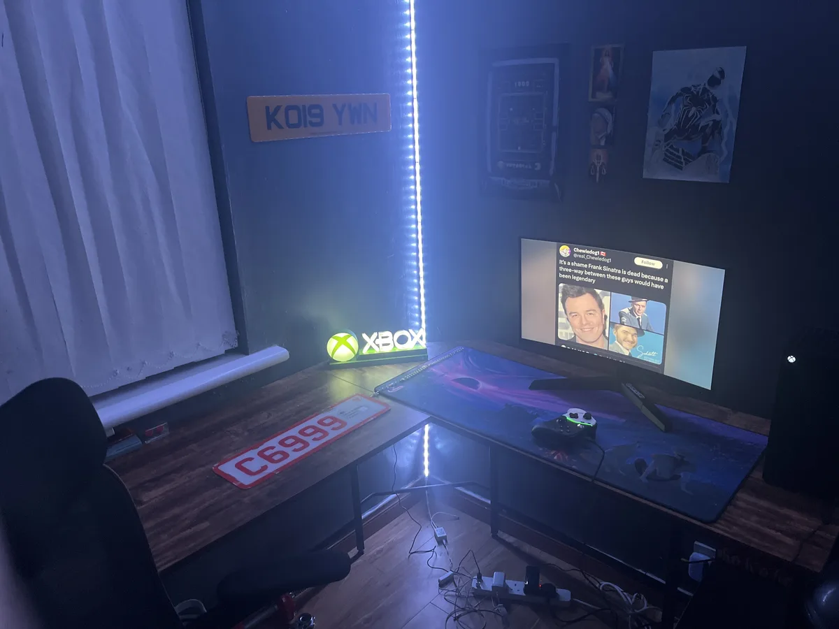 A dimly lit gaming setup with blue LED lighting, featuring an Xbox console, controller, and monitor displaying a tweet from…