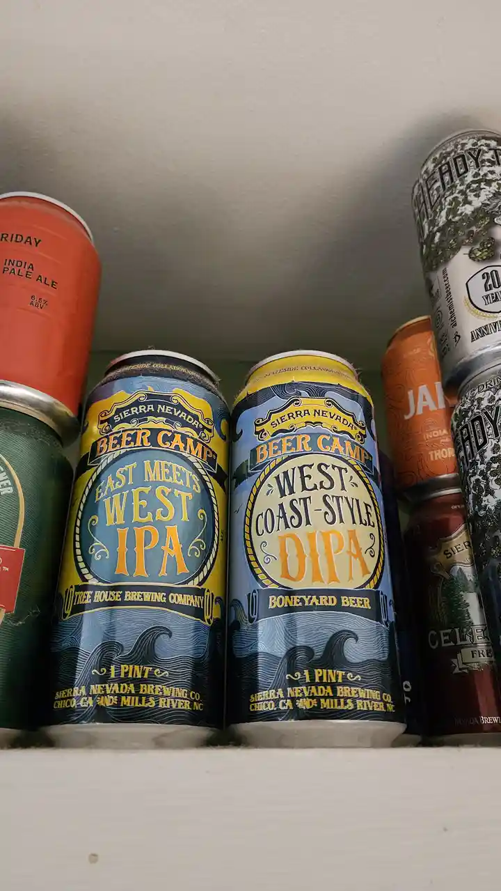 Two Sierra Nevada 'Beer Camp' cans sit side-by-side on a shelf, one labeled 'East Meets West IPA' and the other 'West…