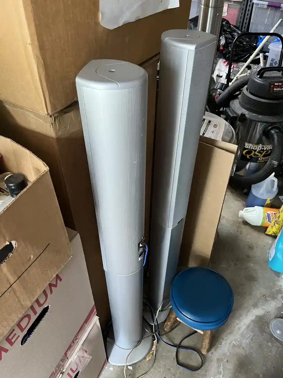 Two tall, silver, cylindrical speakers stand upright in a cluttered garage next to cardboard boxes, a blue stool, and a…