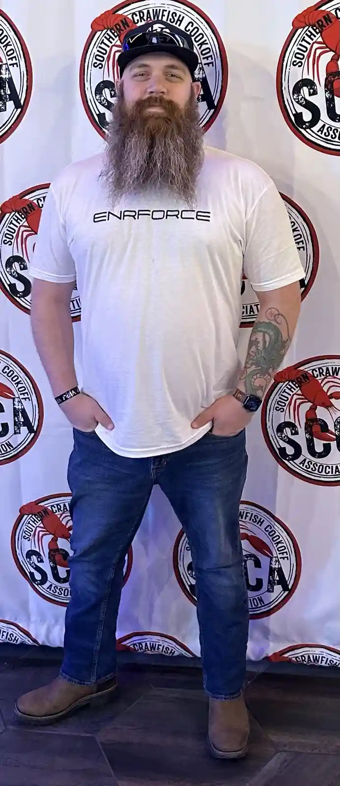 A man with a long beard and a black cap stands confidently with hands in his pockets in front of a backdrop featuring the…