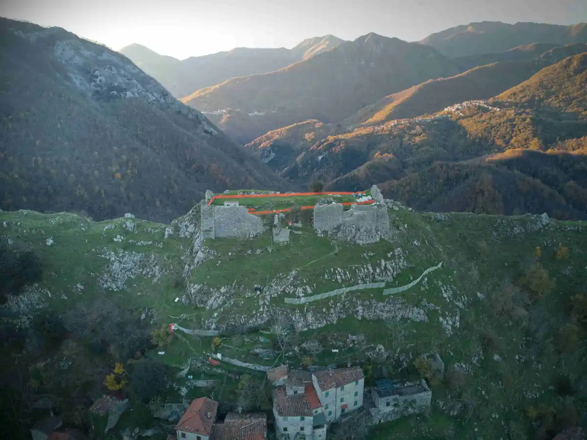 An aerial view shows ancient stone castle ruins perched atop a grassy mountain ridge, surrounded by forested hills and…