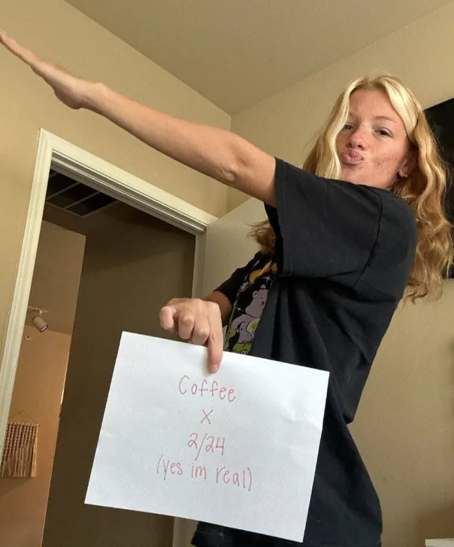 A young woman with blonde hair, wearing a black t-shirt, holds a handwritten sign that reads 'Coffee x 2/24 (yes im real)'…