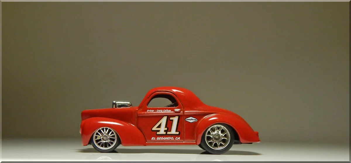 A red die-cast model of a vintage hot rod race car is shown in profile against a plain gray background. The car features the…