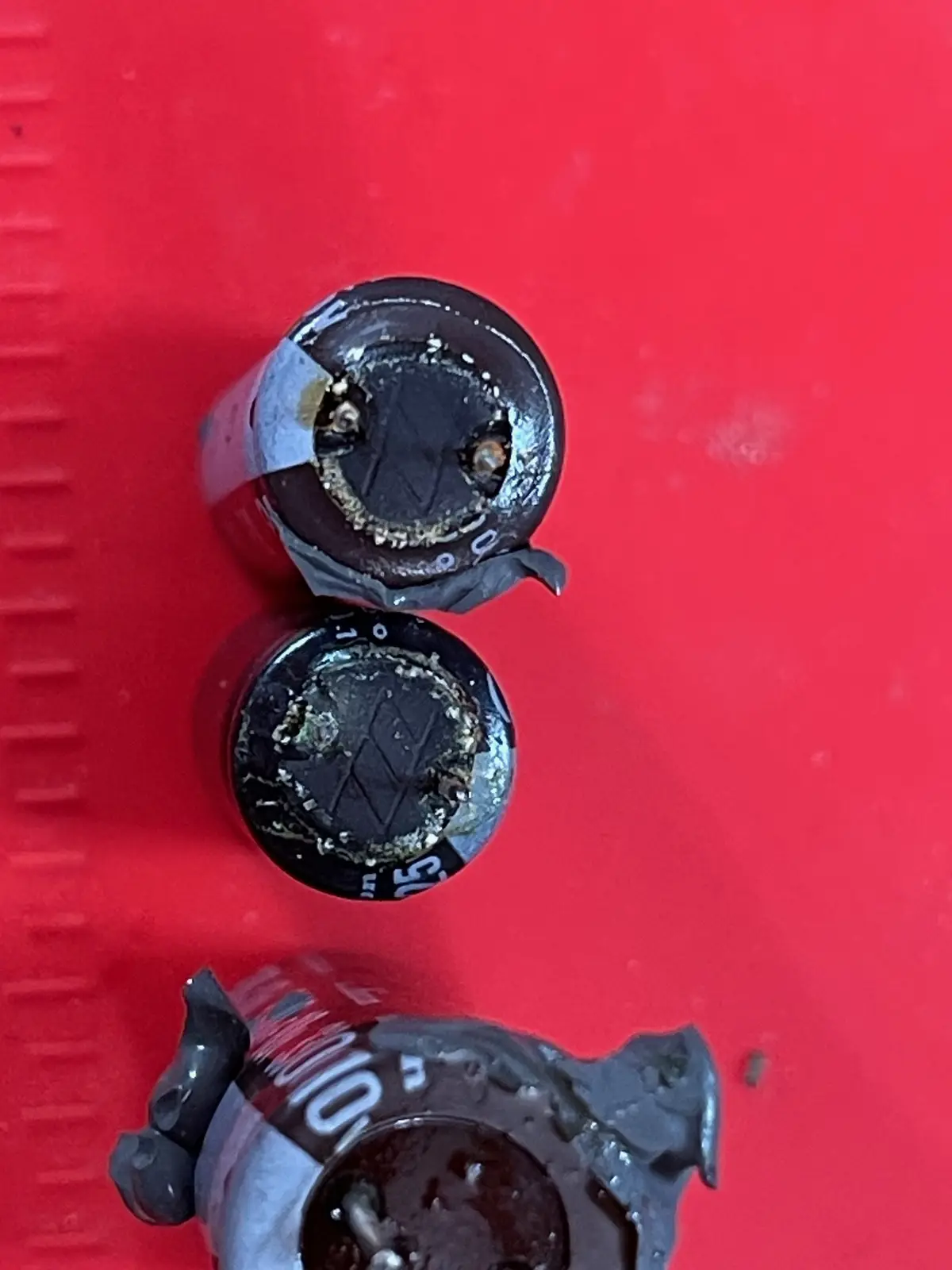 Three corroded, leaking batteries with torn labels sit on a red surface, their ends visibly damaged and leaking a brownish…