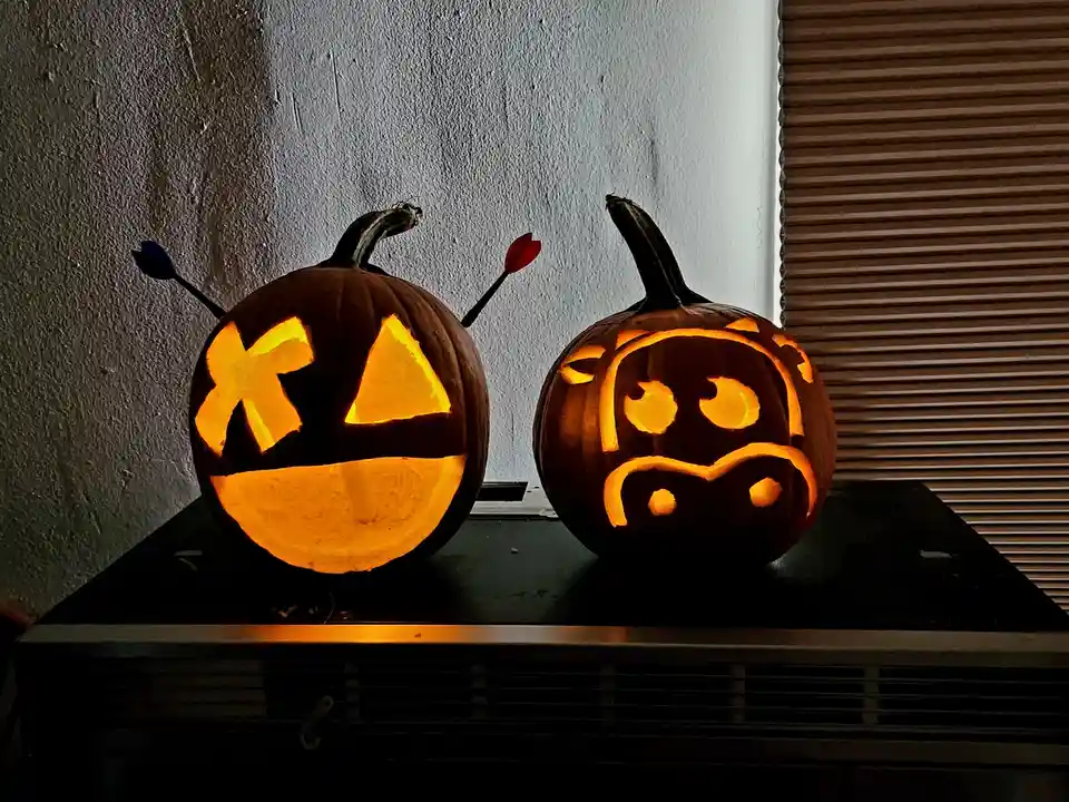 Two carved Halloween pumpkins sit side by side on a dark surface, illuminated from within. The pumpkin on the left has a…