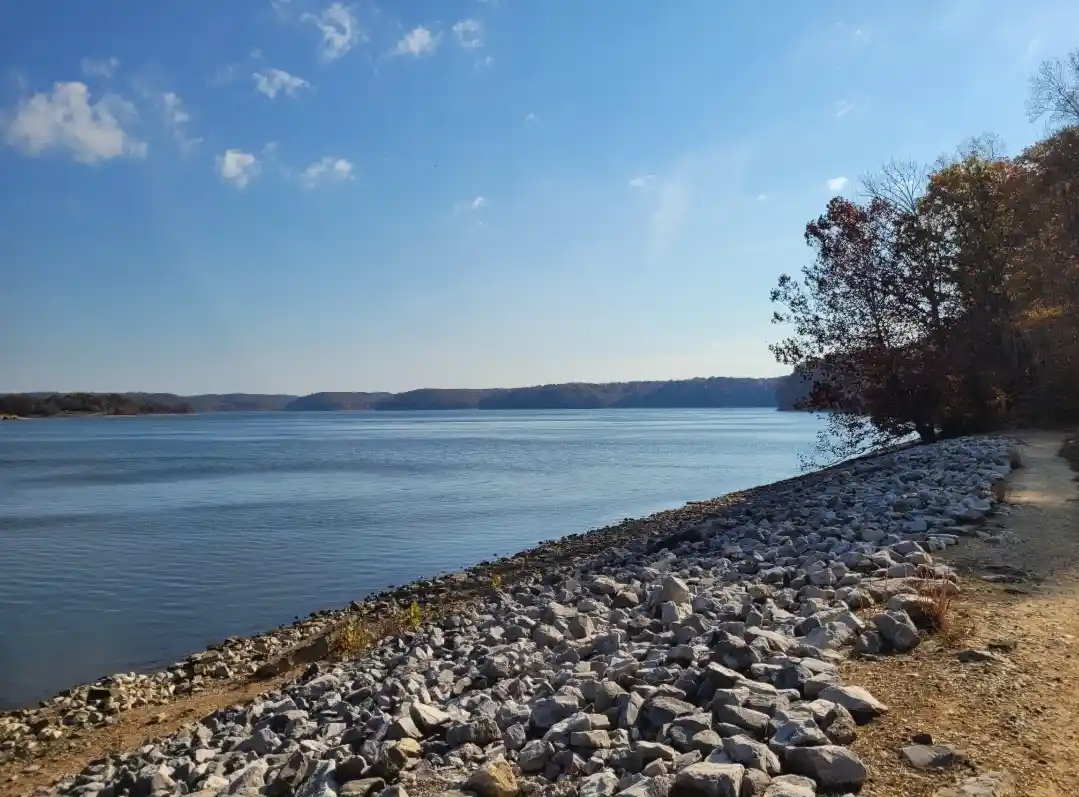 A scenic photo of a calm lake under a blue sky with scattered clouds, bordered by a rocky shoreline and autumn-colored…