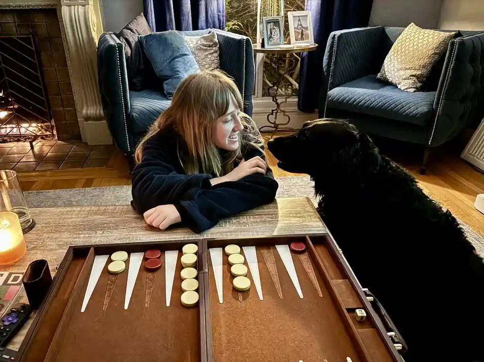 A woman in a dark robe leans over a backgammon board, smiling at a black dog sitting across from her. The dog is looking up…