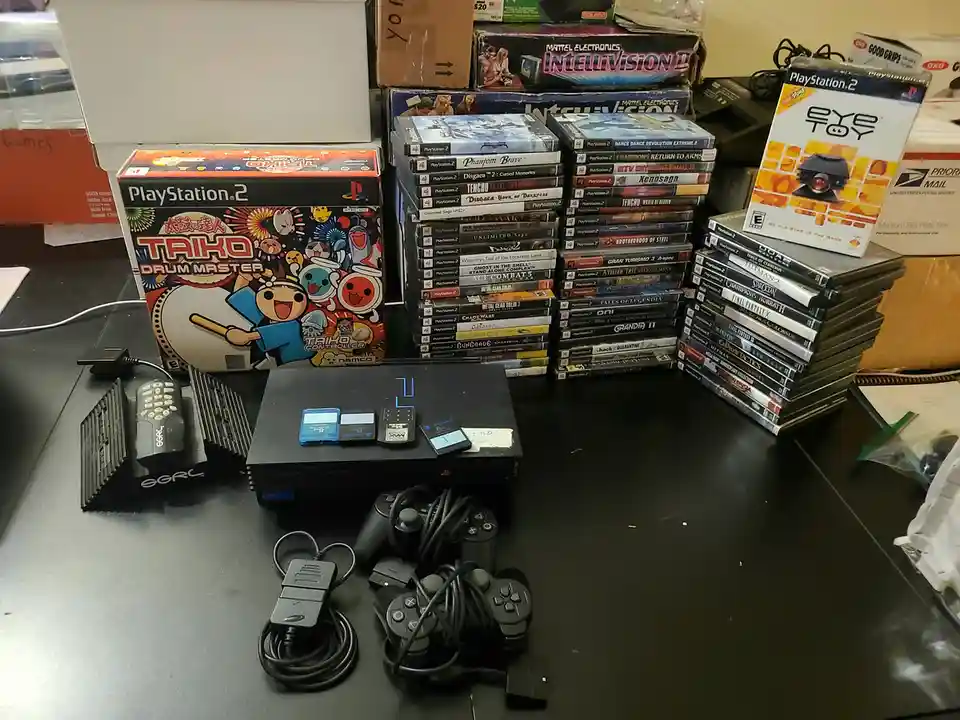 A PlayStation 2 console sits on a desk surrounded by stacks of PlayStation 2 game cases, a 'Taiko Drum Master' game box…