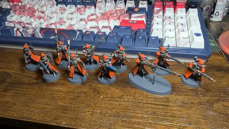 A row of eight painted miniature warhammer 40k space marine figures in orange and grey armor stand on a wooden desk in front…