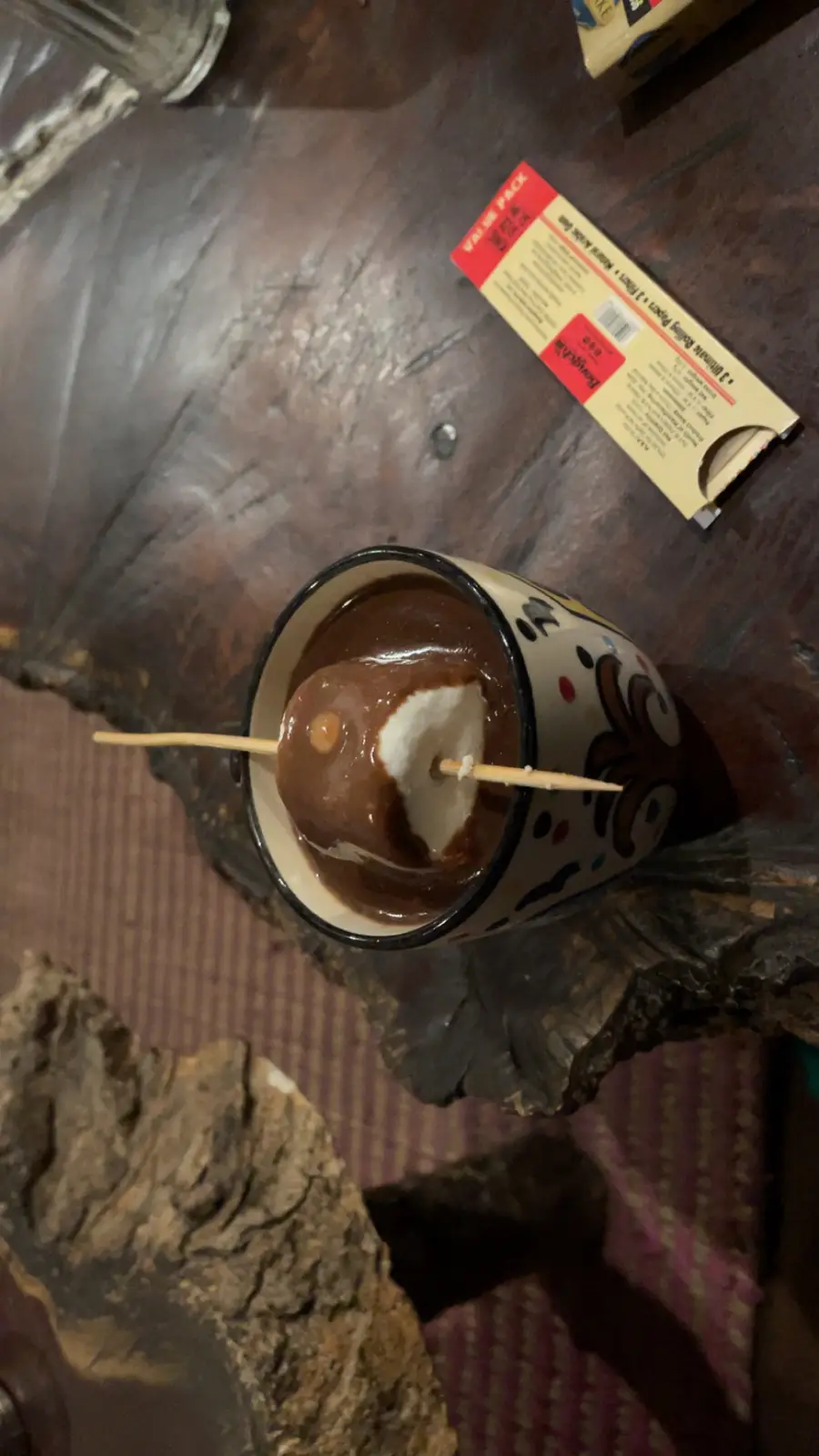A close-up photo shows a marshmallow on a stick partially dipped in melted chocolate inside a patterned mug, with a visible…
