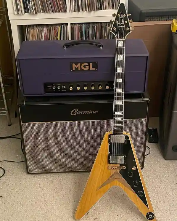 A light-wood, V-shaped electric guitar with a black pickguard and 'Baukler' logo on the headstock stands upright on a…