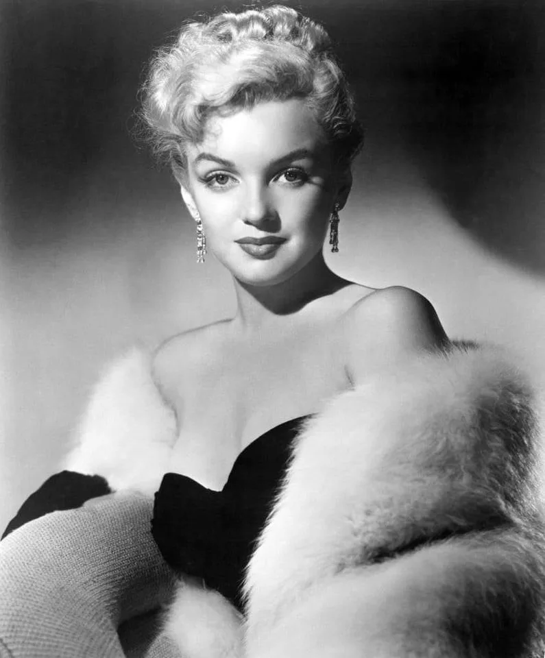A black-and-white portrait of Marilyn Monroe wearing a black dress and white fur stole, looking directly at the camera with…