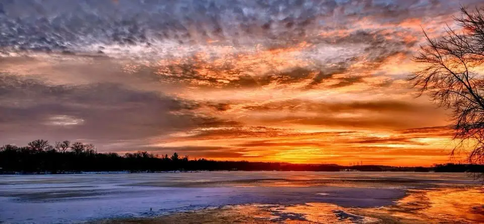 A serene winter sunset over a frozen lake, with vibrant orange and purple clouds reflecting on the icy surface. Bare tree…