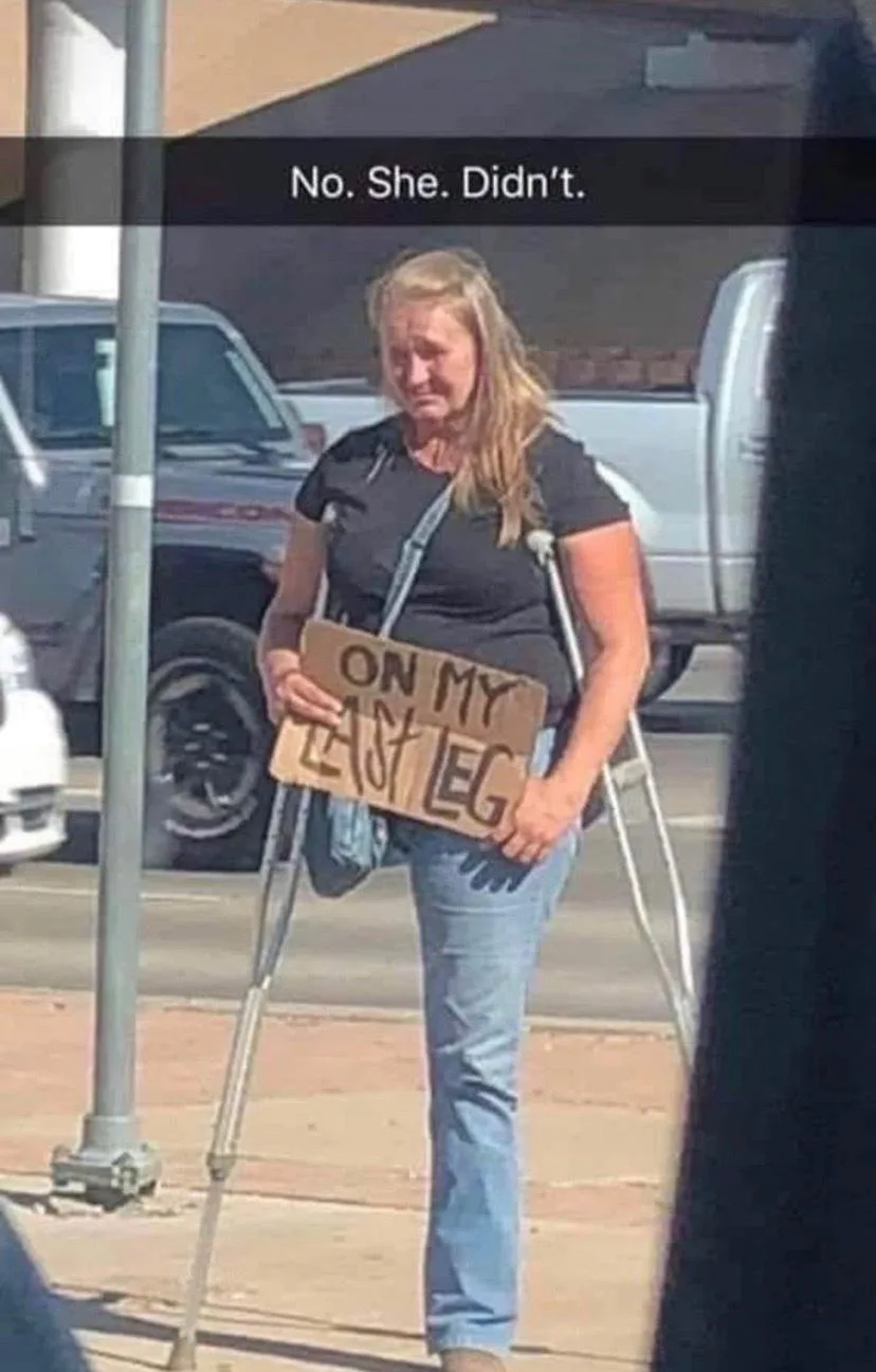A woman with blonde hair, wearing a black t-shirt and jeans, stands on a sidewalk holding a cardboard sign that reads 'ON MY…