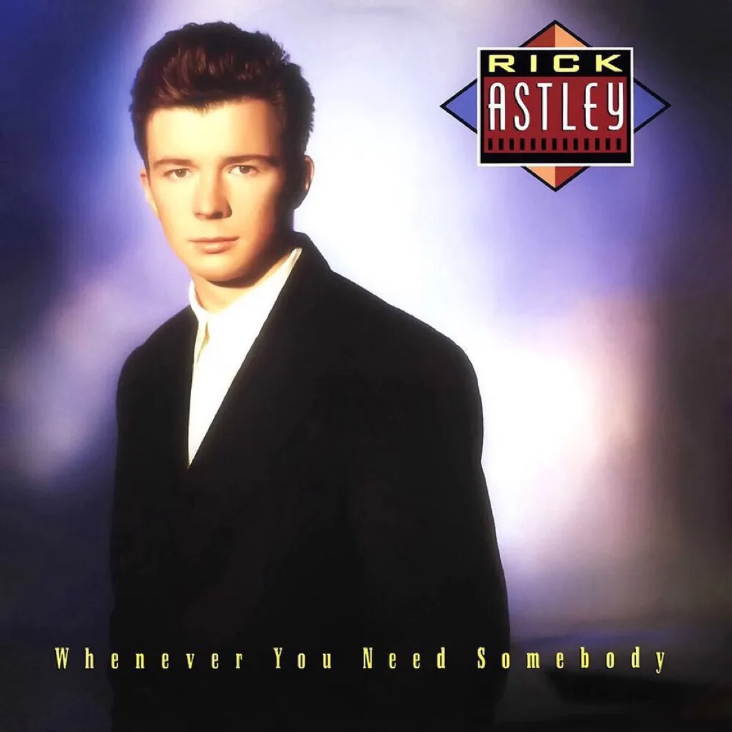 The image shows a young Rick Astley in a black suit and white shirt, looking directly at the camera against a purple and…