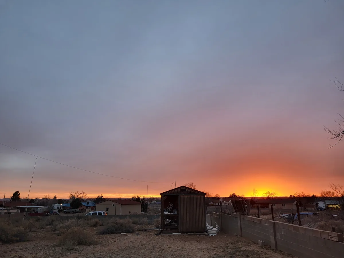 A photograph of a suburban backyard at sunset, featuring a wooden shed with its door open, revealing clutter inside, set…