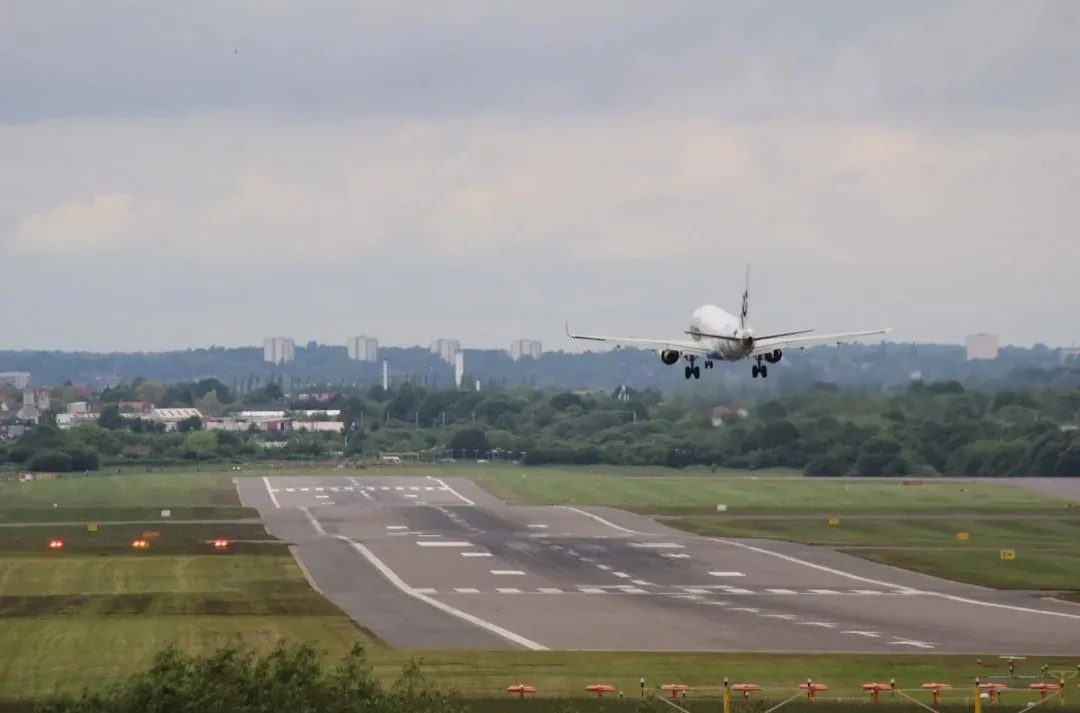 An airplane is captured mid-air, just after taking off from a runway, with a cityscape visible in the background under an…
