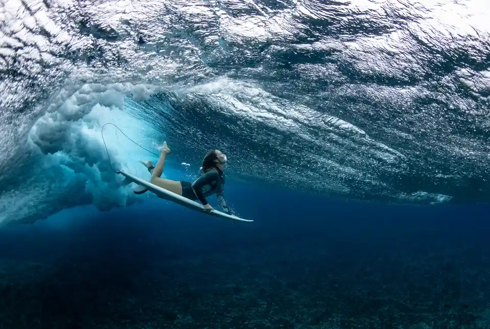 An underwater photo shows a surfer in a wetsuit lying on a surfboard inside a curling ocean wave, looking upward with a…