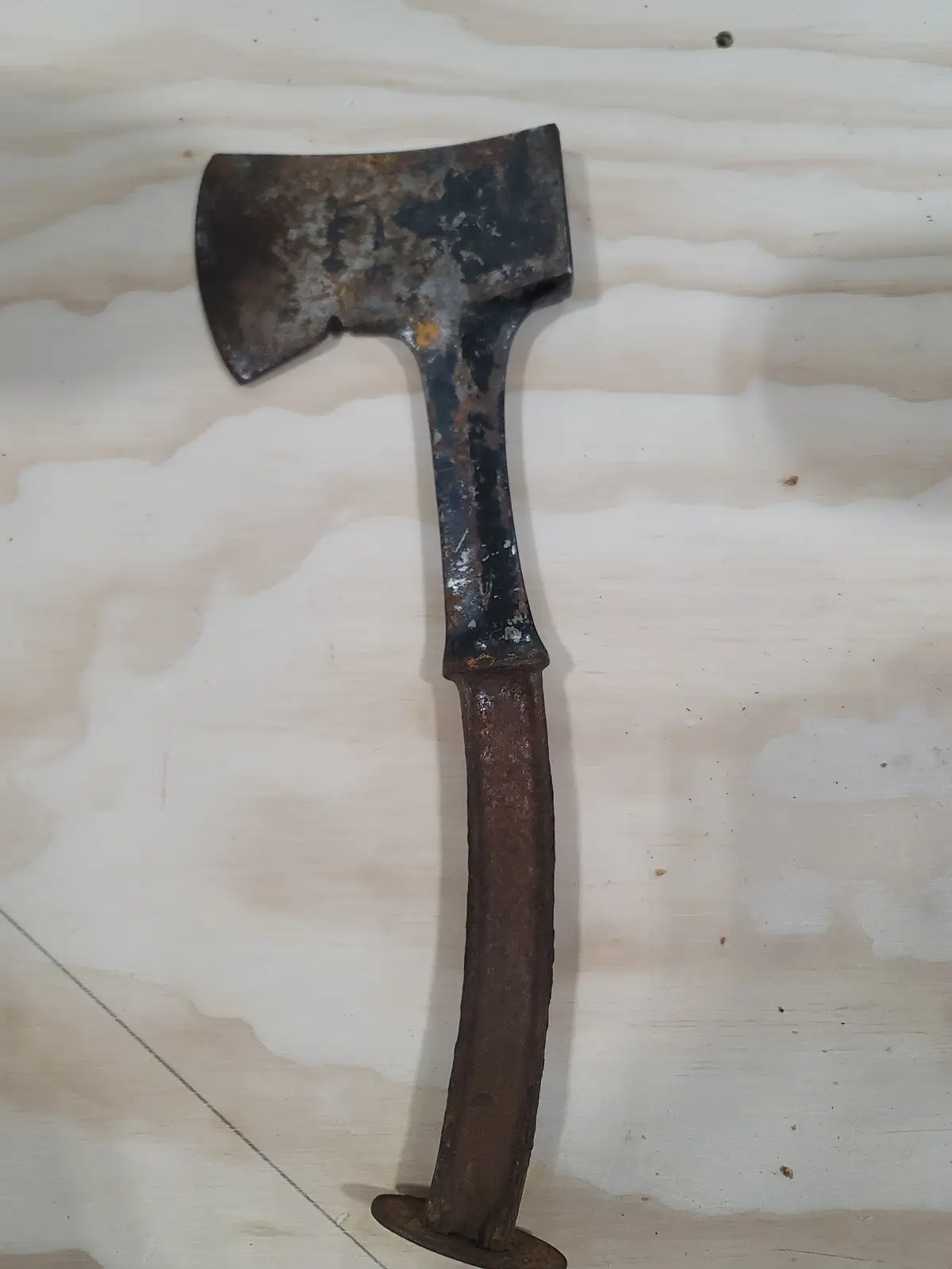 A heavily rusted, old axe with a thick, blunt head and a long, corroded metal handle lies on a light-colored plywood…