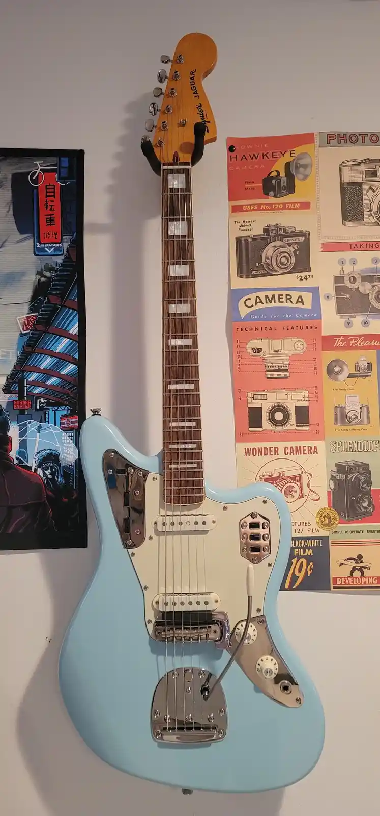 A light blue Fender Jaguar electric guitar hangs on a wall next to vintage camera advertisement posters. The guitar's body…