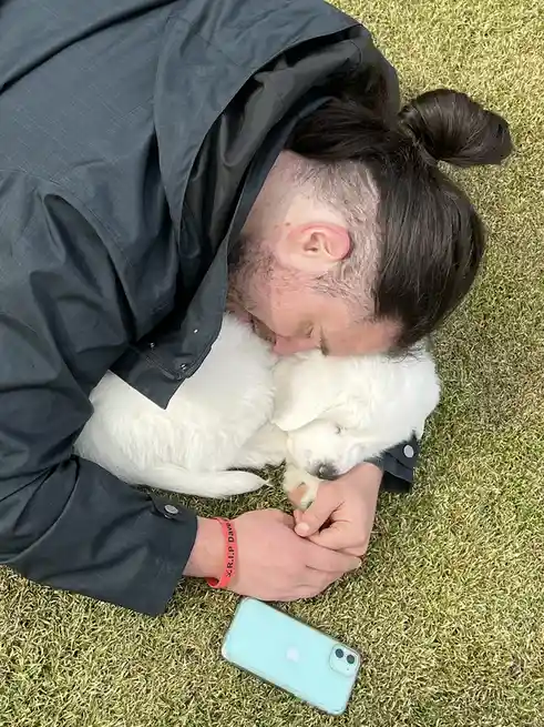 A man in a dark jacket lies on grass, hugging a white fluffy puppy, with a red wristband reading 'R.I.P. Dave' and a light…