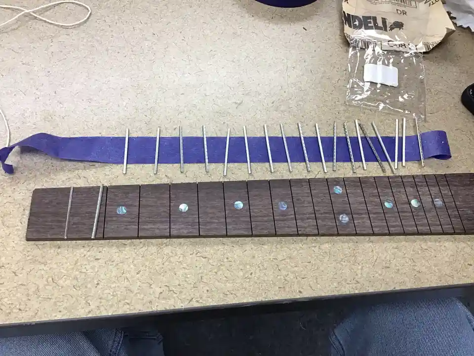 A wooden guitar fretboard with abalone inlays lies on a speckled countertop, with a long purple ribbon and numerous metal…
