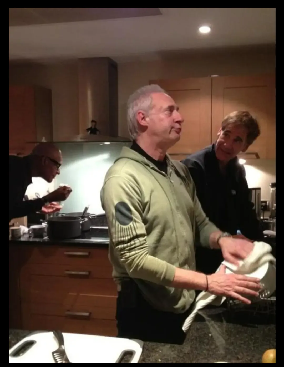 Three men are in a kitchen; one in a green hoodie dries a dish while looking upward with a resigned expression, another man…