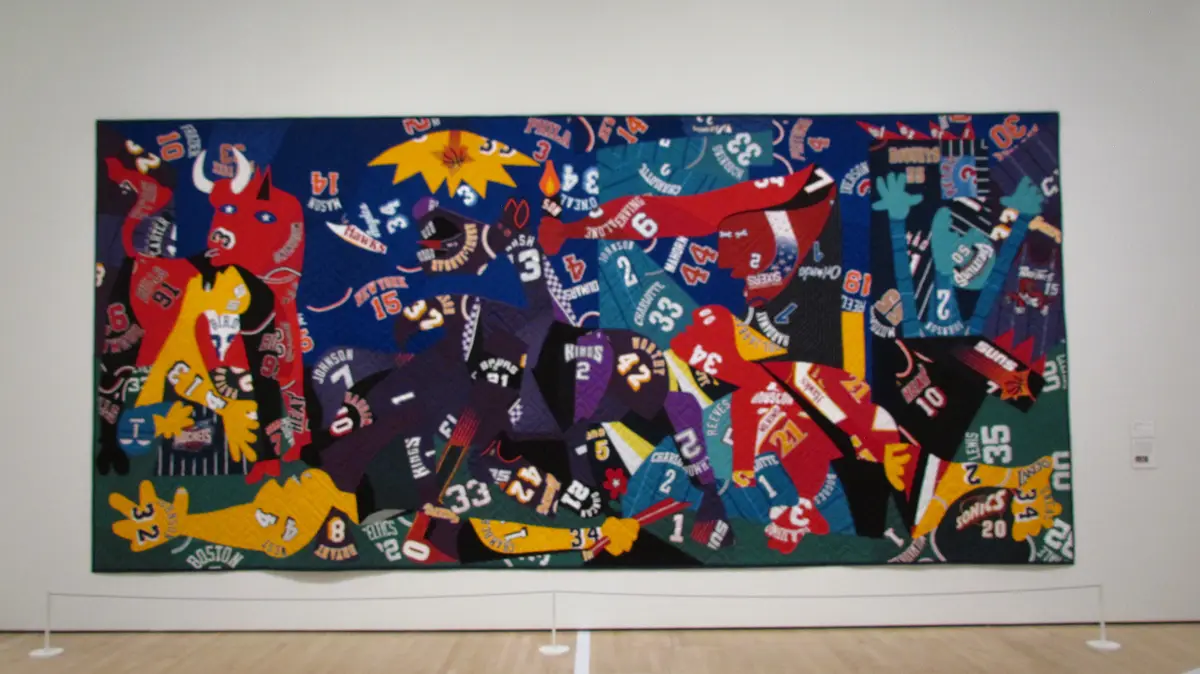 A large, colorful quilt hangs on a gallery wall, constructed from numerous cut pieces of old basketball jerseys featuring…