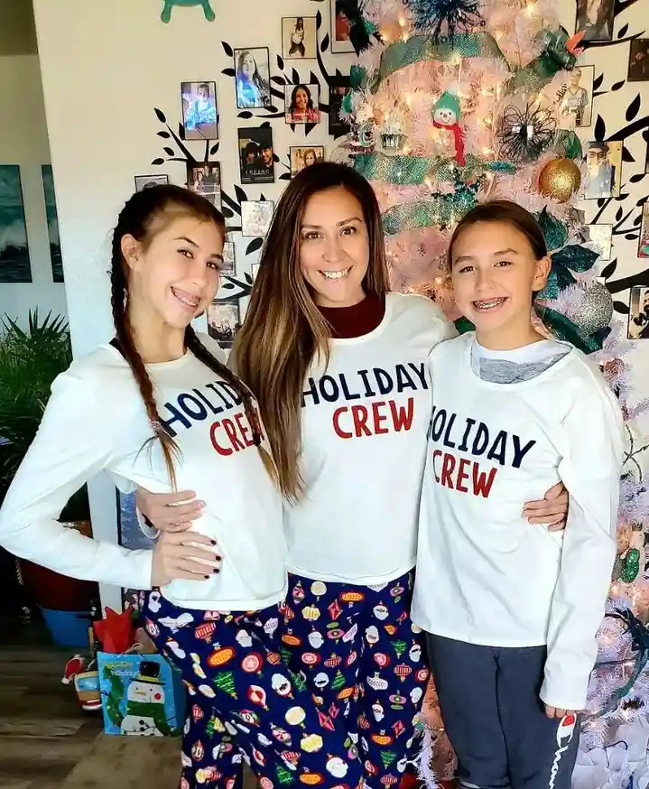 Three people, likely a mother and her two daughters, pose together in front of a decorated pink Christmas tree. They are all…