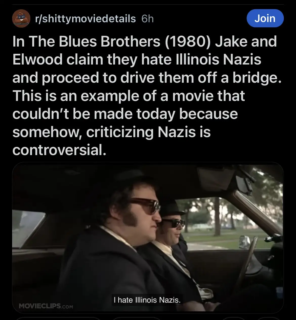 Two men in black suits and sunglasses sit in a car, one driving, the other in the passenger seat. The image is from the 1980…
