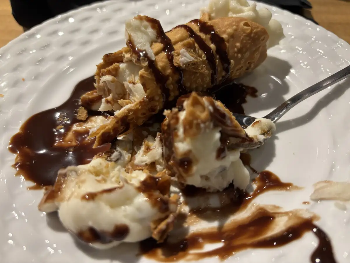 A partially eaten cannoli on a white plate, with its shell broken open to reveal creamy filling, drizzled with chocolate…
