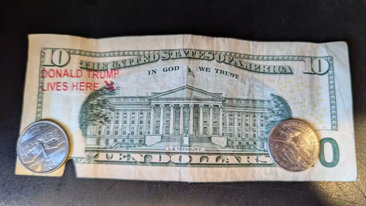A crumpled ten-dollar U.S. bill with red text overlaid that reads 'DONALD TRUMP LIVES HERE', placed on a dark surface with a…