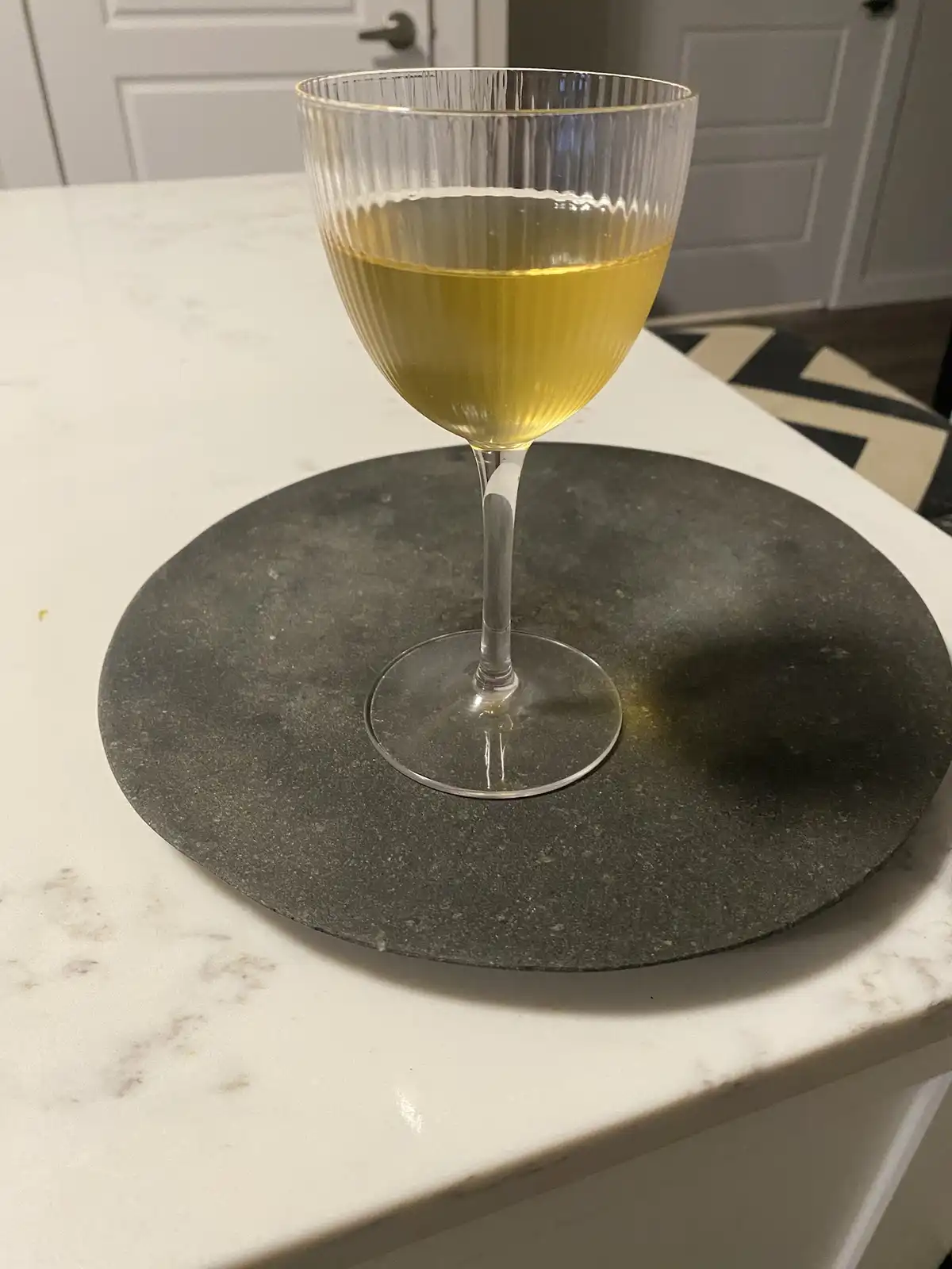 A clear, ribbed wine glass filled with pale yellow liquid sits on a dark circular coaster atop a white marble countertop. In…