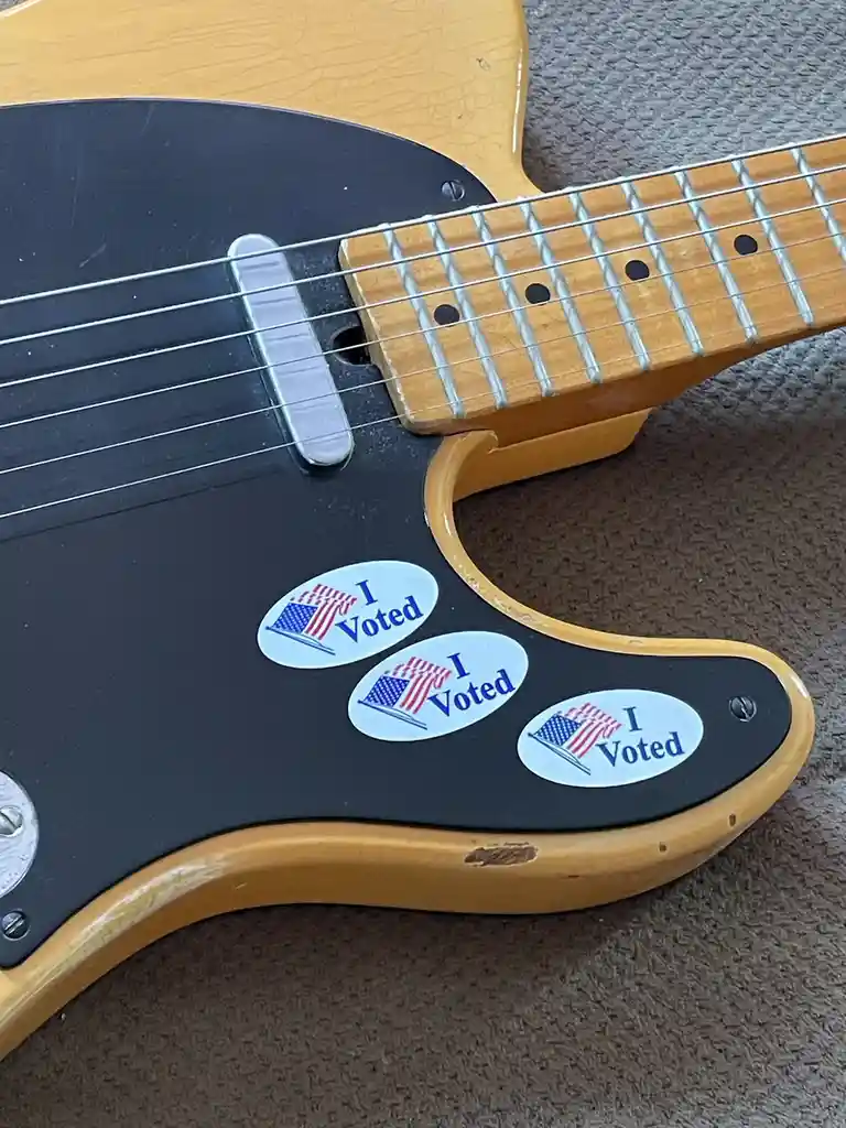 A close-up of a light-wood electric guitar's body, showing three "I Voted" stickers with American flag designs stuck on the…