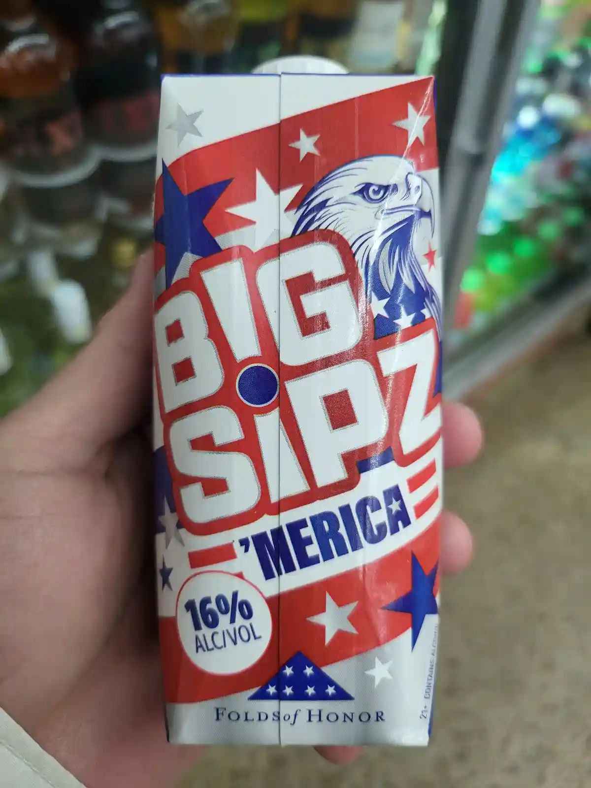 A hand holds a patriotic-themed carton of alcoholic beverage labeled 'BIG SIPZ 'MERICA' with a 16% alcohol content…