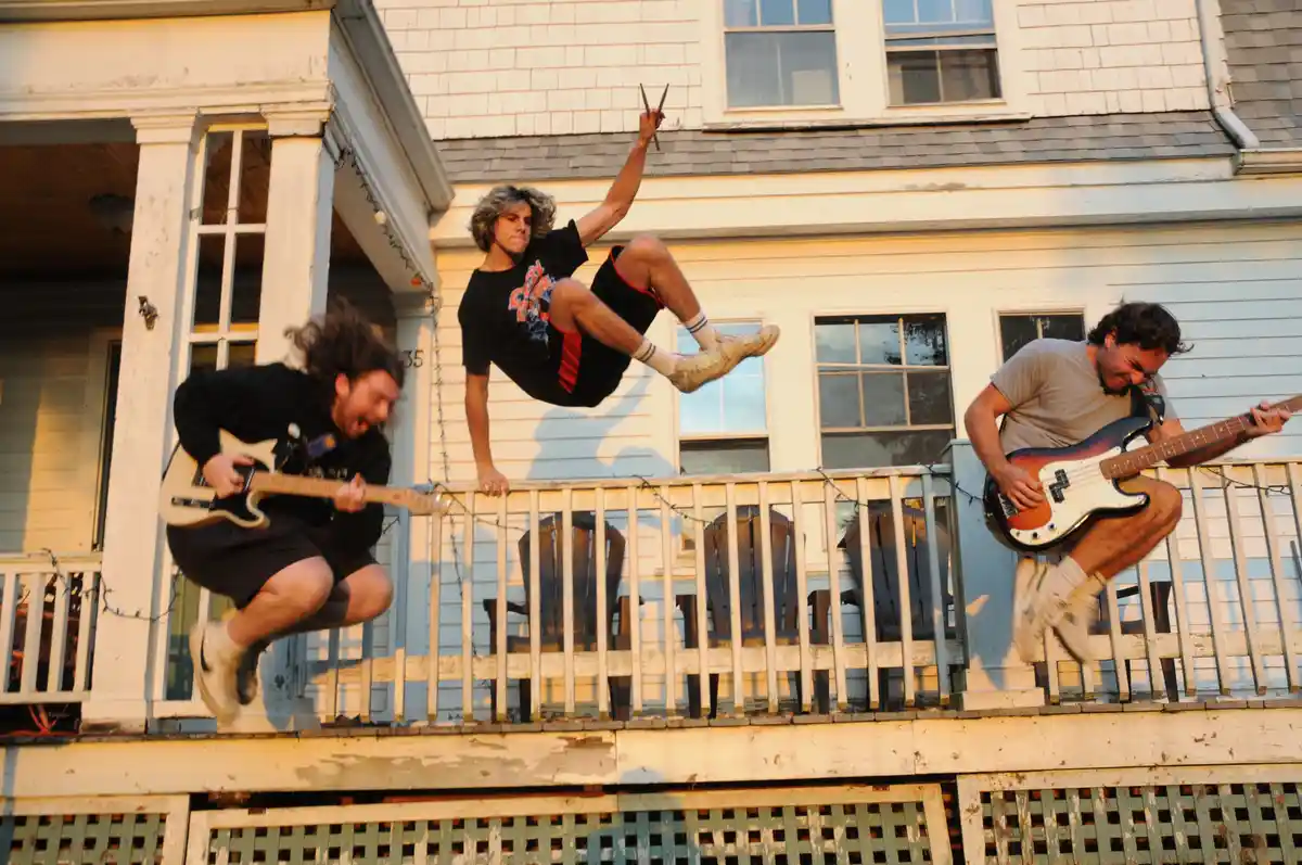 Three men are captured mid-air, seemingly jumping off a porch while playing instruments: one with a guitar on the left, one…