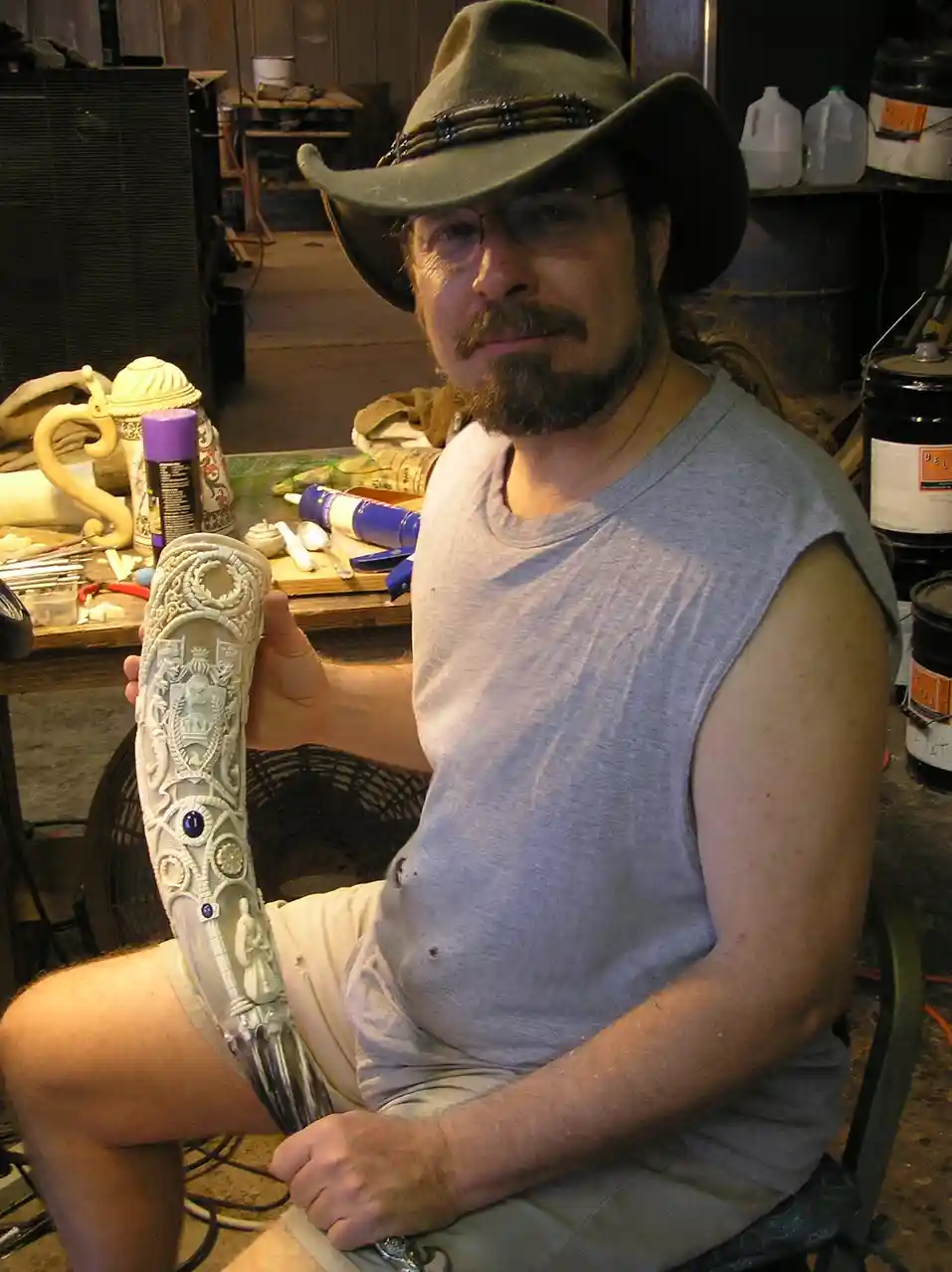 A man with a beard, glasses, and a cowboy hat sits in a workshop holding an ornate, highly detailed sword hilt he has…