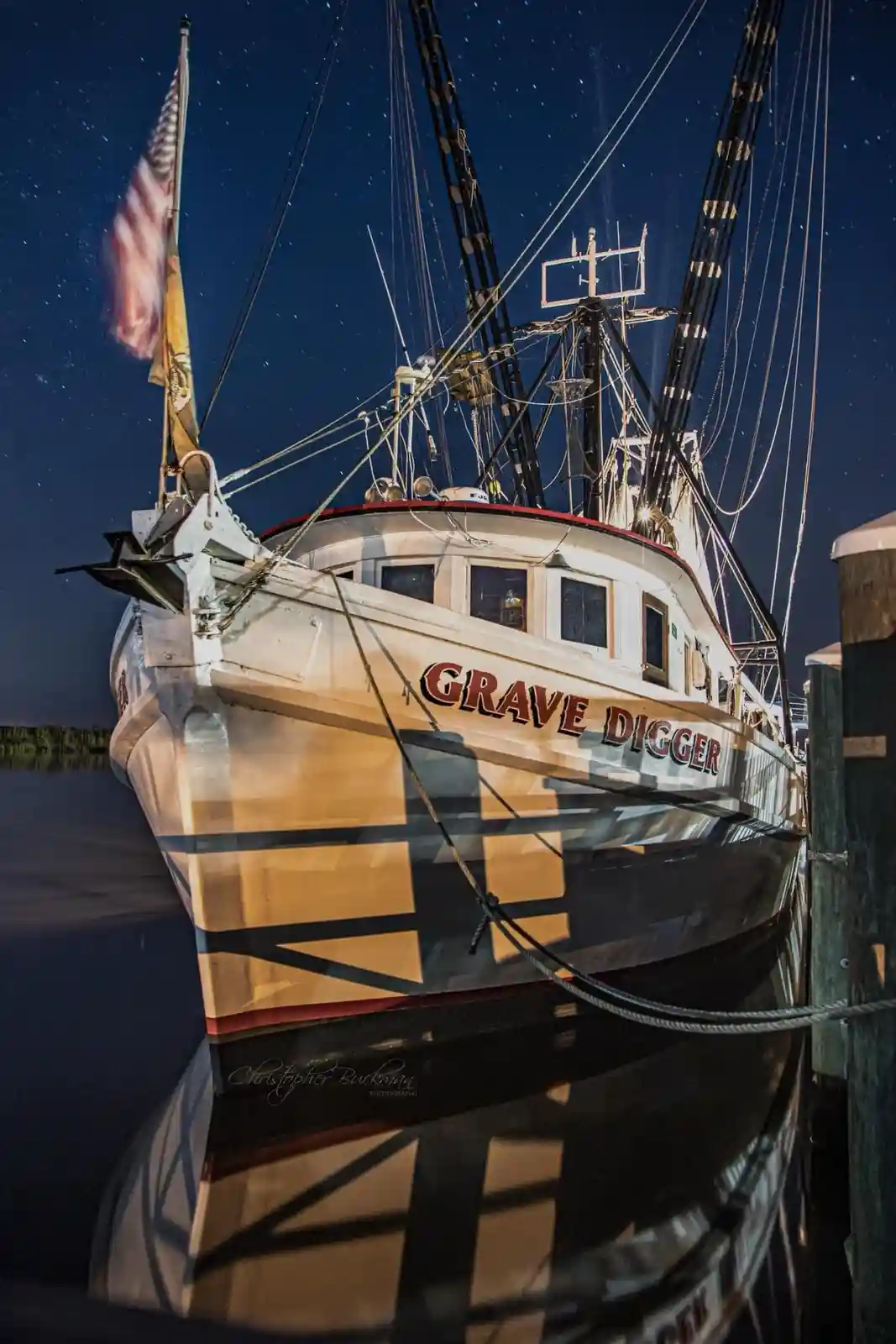 A nighttime photograph of a shrimp boat named 'GRAVE DIGGER' docked at a pier under a starry sky, with its name prominently…